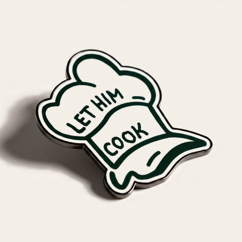 The Chef golf ball marker, a white chef hat outlined in forest green reading LET HIM COOK, photographed at an angle.