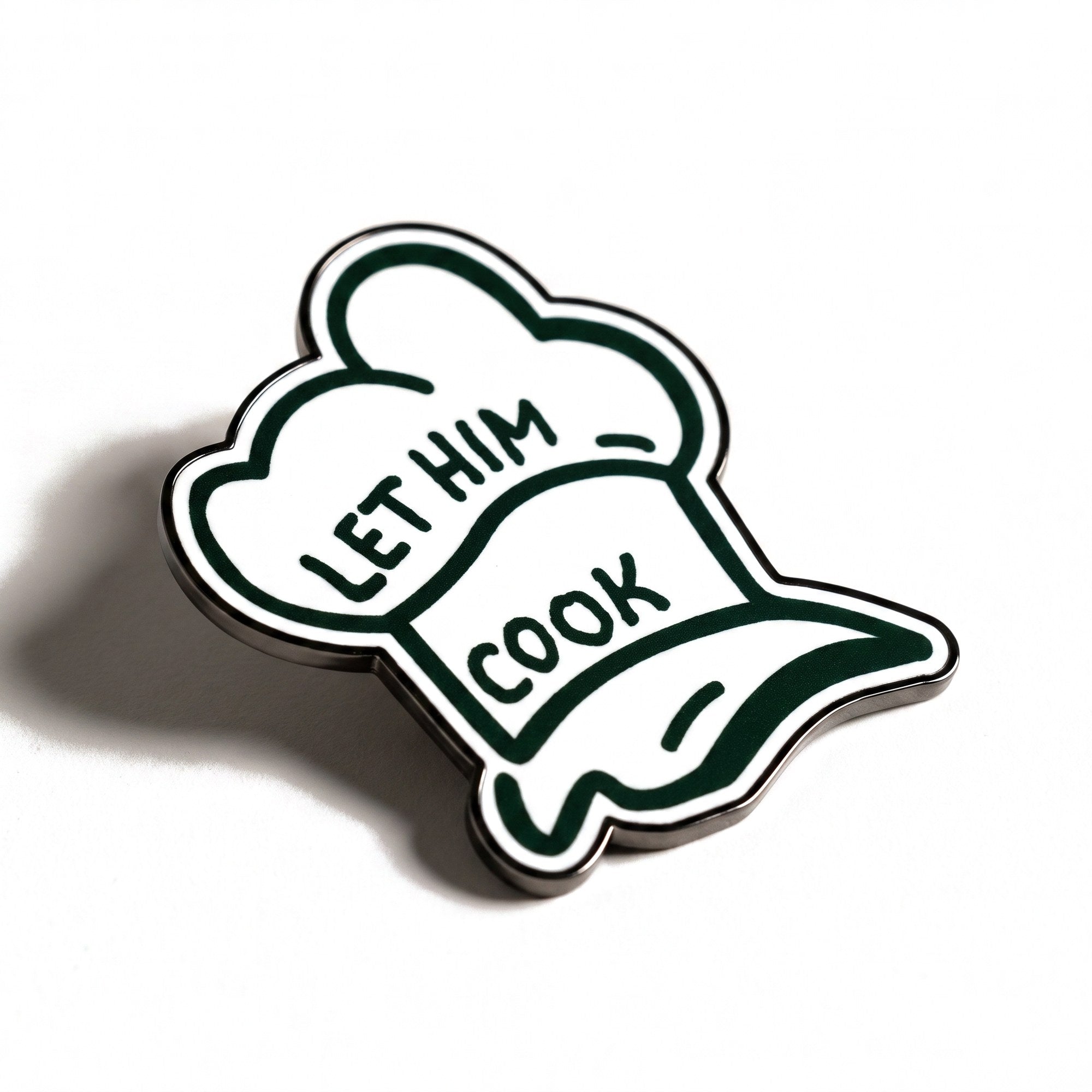 The Chef golf ball marker, a white chef hat outlined in forest green reading LET HIM COOK, photographed at an angle.