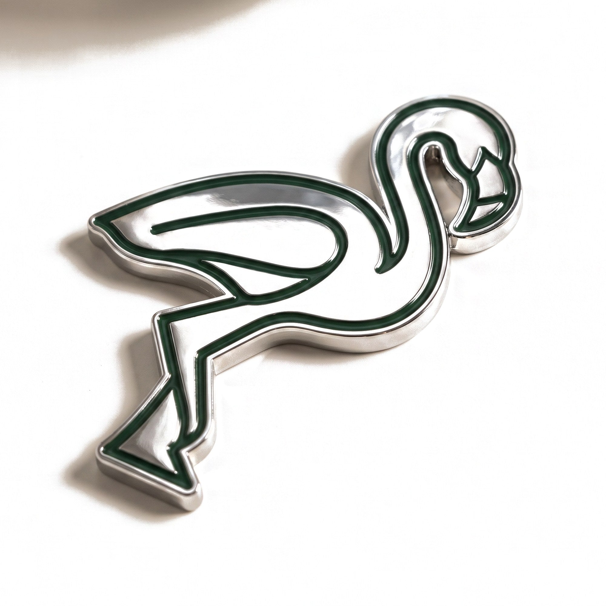The Flamingo golf ball marker, a polished chrome flamingo silhouette with recessed forest green line work, photographed at an angle.