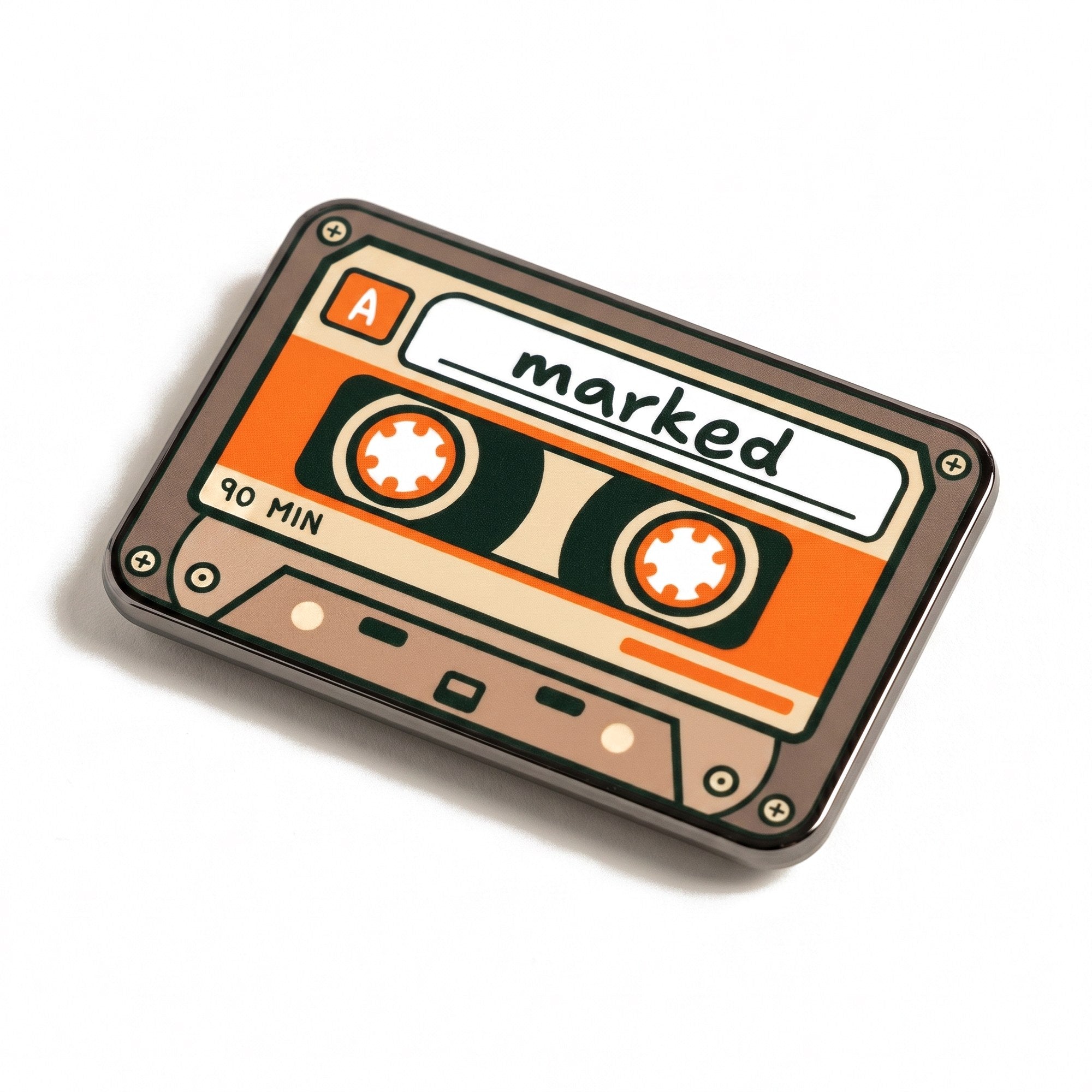 The Soundtrack golf ball marker, a taupe and orange cassette tape with marked handwritten on the label, photographed at an angle.