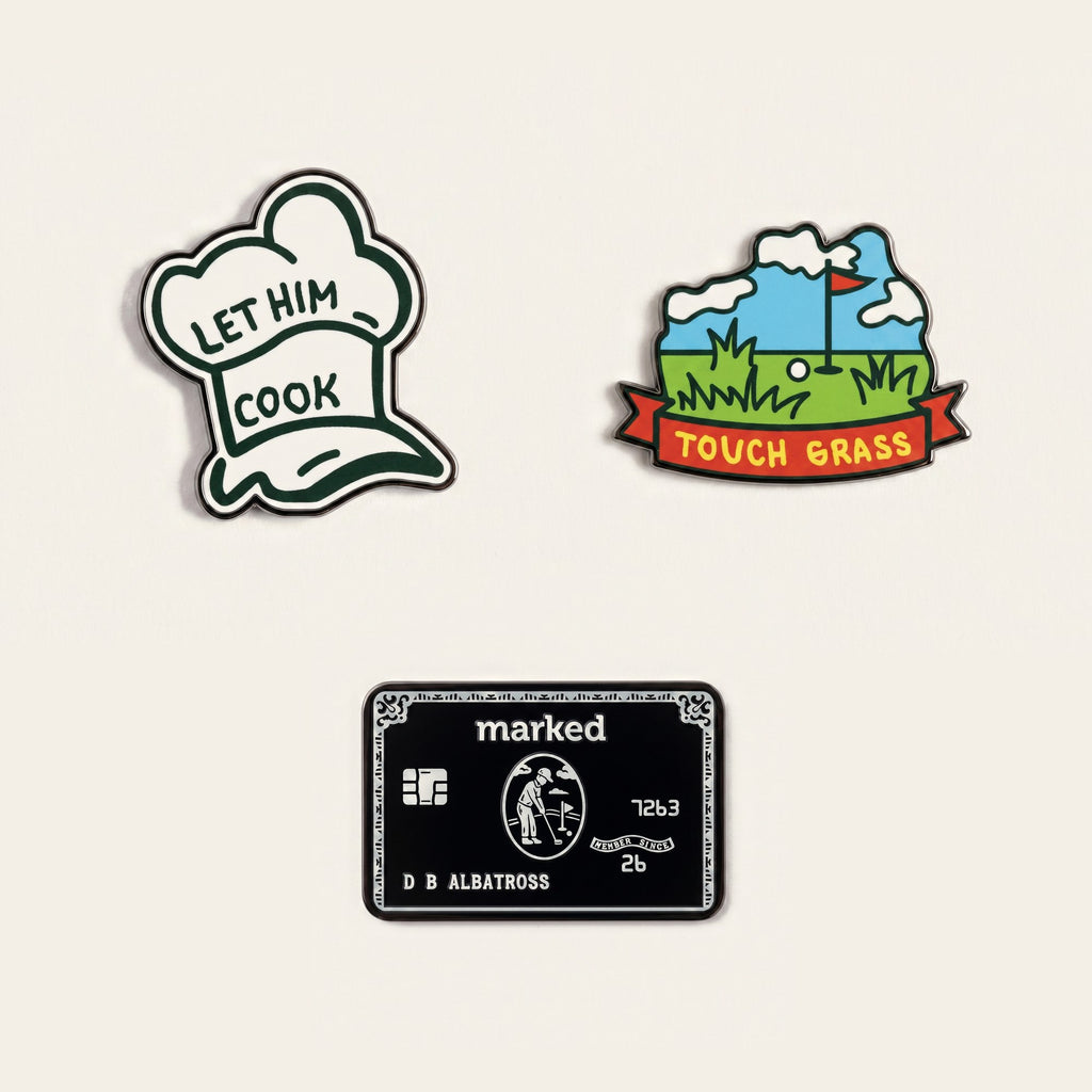 The Group Chat ball marker set photographed flat from above on a cream background: a white chef hat, a fairway scene reading TOUCH GRASS, a black charge card, and the chrome Monogram Signature marker.