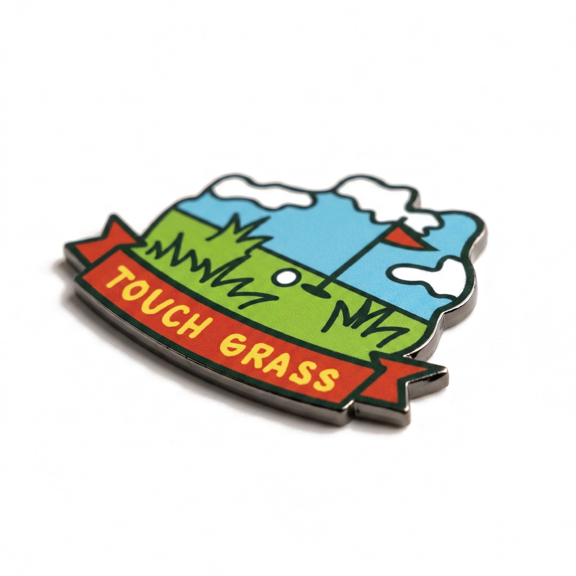 The Touch Grass golf ball marker, a die-cut badge showing a green fairway under a blue sky with a red banner reading TOUCH GRASS, photographed at an angle.
